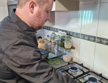 A man with short hair in a dark shirt is cooking, adjusting knobs on a gas stove. In the background, jars of various dried goods and spices are neatly arranged on the kitchen countertop. The kitchen has white tiled walls with a decorative border.