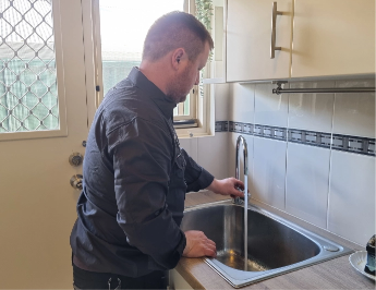 A person with short hair, wearing a dark shirt, stands at a kitchen sink with water running from the tap. The kitchen has white tiles and light-colored cabinets. In the background, there is a door with a mesh screen.