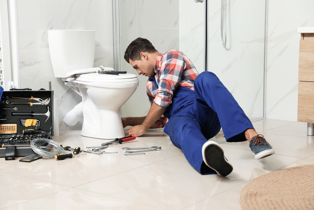 A plumber in blue overalls kneels on a bathroom floor next to a toilet, delivering expert commercial plumbing services. He uses a tool for repairs while an open toolbox brimming with various tools sits nearby. The bathroom is adorned with white tiles and boasts a wooden cabinet.