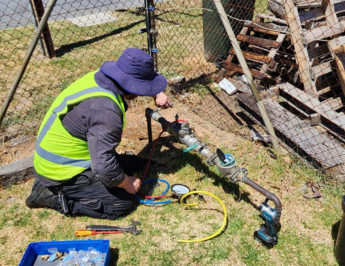 A worker in a neon yellow safety vest and blue hat kneels on grass, adjusting a water meter connected to a pipe. Tools and equipment are scattered nearby, with a chain-link fence and wooden pallets in the background.