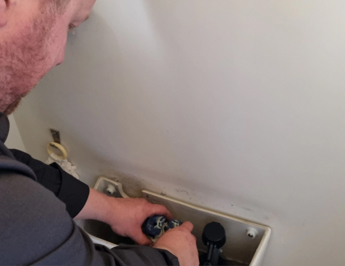 A person with short hair is fixing a toilet tank, adjusting the components inside. The individual is wearing a dark-colored shirt, and their hands are positioned inside the tank beside the fill valve. The wall behind the toilet is plain white.