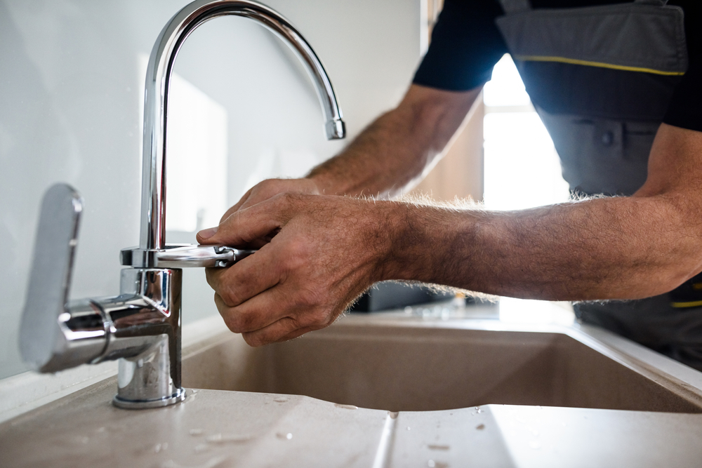 A person wearing a dark shirt and gray overalls is repairing a silver kitchen faucet over a beige sink, expertly adjusting the faucet handle to stop the drip. Water droplets glisten on the sink surface—a reminder that sometimes it's best to simply call a plumber for a leaking tap.