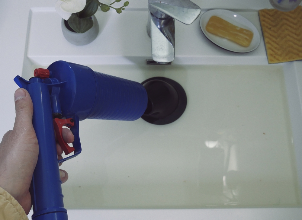 Using a blue plunger gun, a person tackles a sink filled with water. Nearby, a chrome faucet gleams, juxtaposed by a bar of soap on a small dish and a yellow washcloth. As the potted plant adds charm, remember to know when to call a plumber for that pesky leaking tap.