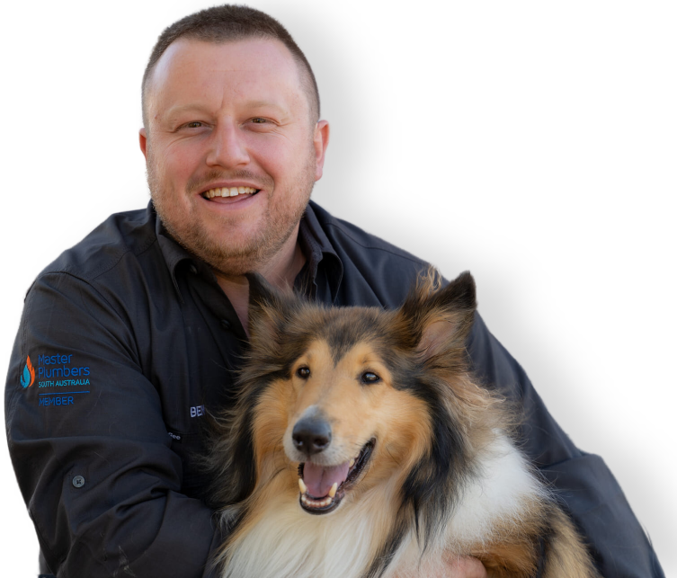 A man in a black shirt with an embroidered logo smiles while holding a happy, long-haired dog. The man has short hair and facial stubble. The dog appears joyful with its mouth open and ears perked up.