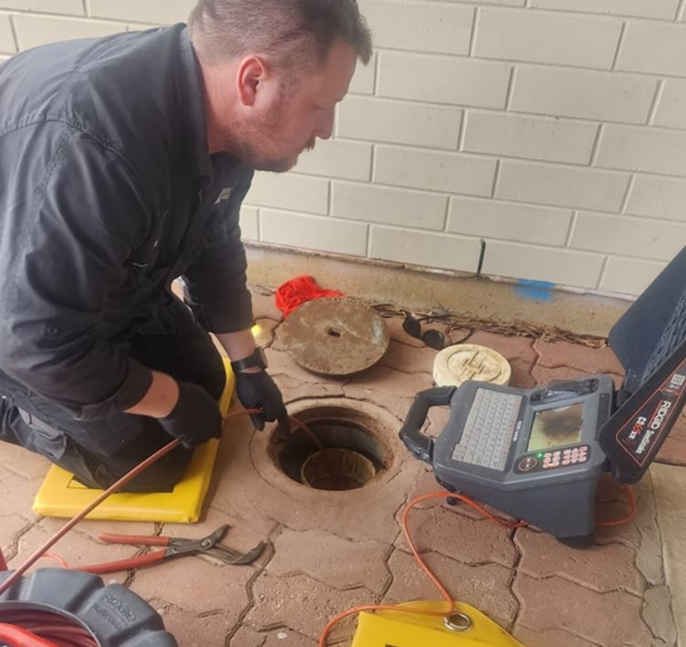 A technician in protective clothing kneels on a paving stone floor, operating sewer inspection equipment. He uses a monitor and cable to examine a manhole. Nearby are tools, including pliers and a yellow case.