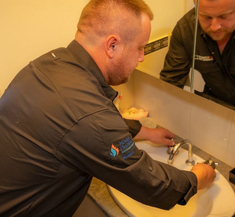 A person in a dark uniform shirt is fixing a faucet in a bathroom. They are leaning over a sink with hands on the tap, reflected partially in the mirror above the sink. Various toiletries are visible on the counter.