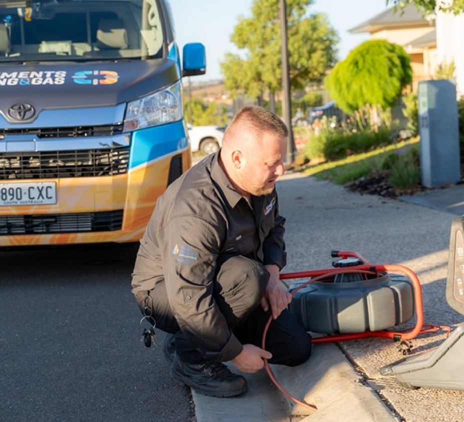 A man in a dark uniform kneels on a sidewalk, inspecting equipment with orange cables. A van with logos and branding is parked behind him on the street, and a residential area with trees and houses is in the background.