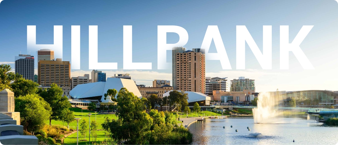 Skyline view of a city with modern buildings and lush green park in the foreground. A large fountain is spraying water in a lake, and the word "HILLBANK" is prominently displayed across the sky.