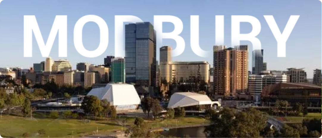 A city skyline with numerous modern buildings under a clear blue sky. The word "MODBURY" is prominently displayed across the top of the image. Green trees and a river are visible in the foreground.