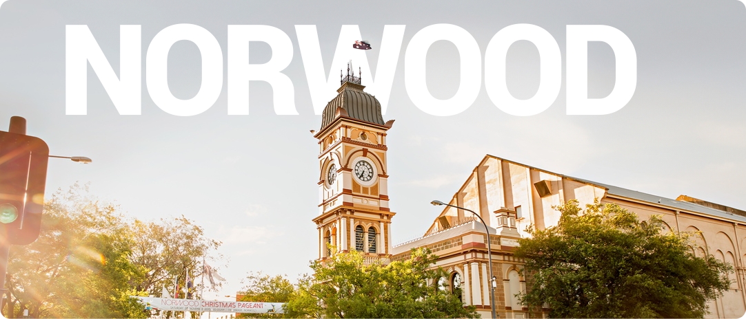 A historic clock tower surrounded by trees stands under a clear sky, with the word "Norwood" prominently displayed in bold white letters across the top of the image.