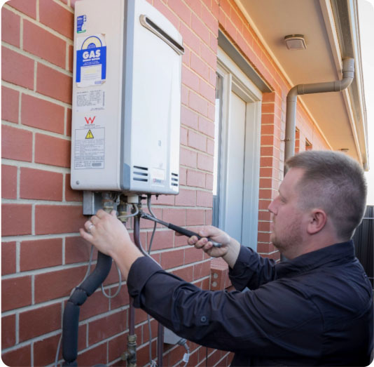 Image 137 A man in a dark shirt is servicing or inspecting a gas water heater mounted on the exterior brick wall of a house. He is using a tool to adjust or check the pipes connected to the unit.