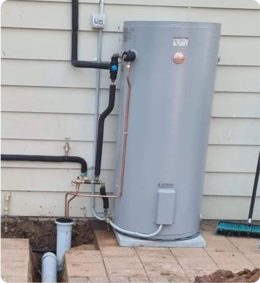 Image 138 A gray water heater is installed outdoors on a concrete pad next to a beige building. Pipes connect the heater to plumbing in the ground. A blue broom is lying nearby on the right.