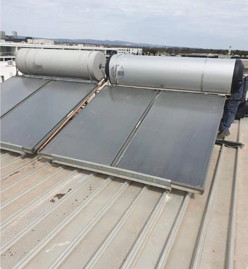 Two solar water heaters with large metal tanks and flat solar panels are installed on a sloped rooftop. The sky is cloudy, and buildings are visible in the background.