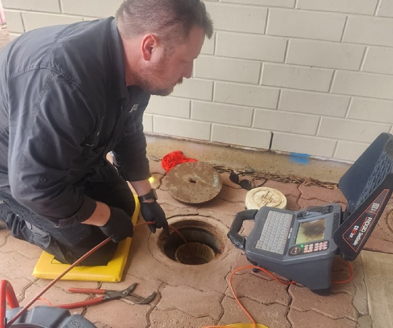 A man wearing black work clothes and gloves inspects a drain using a cable and a sewer inspection camera, with the monitor displaying footage next to him on the ground. Tools are scattered nearby.
