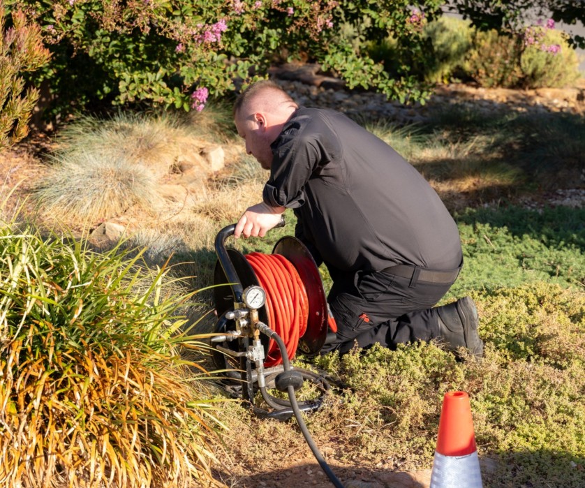 commercial plumber A person in black clothing kneels on the ground outdoors, operating a hose reel near green and brown plants. A traffic cone is visible in the foreground, and trees and bushes are in the background.