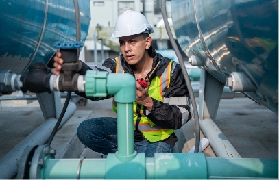 industrial-engineer-checking-equipment-with-tool-2025-03-06-15-59-38-utc A worker wearing a hard hat, safety vest, and jeans inspects and adjusts a valve on industrial pipes between large blue tanks in an industrial facility.
