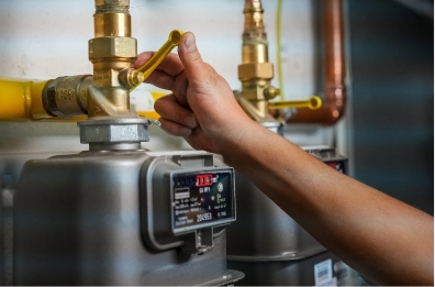 person-adjusting-a-gas-meter-to-reduce-gas-usage-2025-02-09-10-40-51-utc A person’s hand is turning a yellow valve handle on a gas meter connected to yellow pipes, with another meter visible in the background.