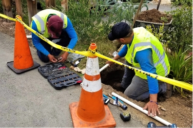Two workers in safety vests repair pipes in a small pit beside a road, surrounded by caution tape and traffic cones. Various tools are laid out on the ground next to them.
