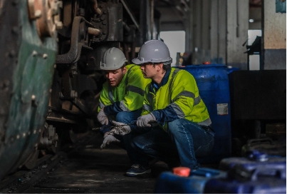 rail-technicians-identify-repair-engine-issues-p-2025-01-07-23-12-05-utc Two workers in safety helmets and high-visibility jackets crouch next to machinery in an industrial setting, inspecting or repairing equipment. Blue containers are visible in the foreground.