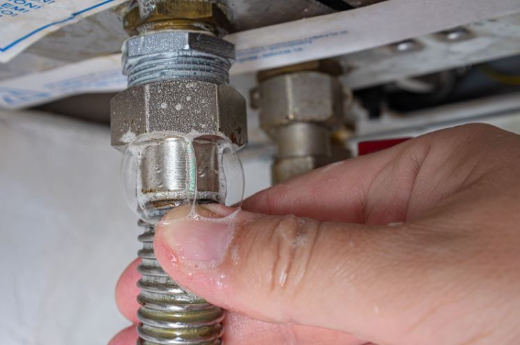 A hand holding a soapy hose under a metal pipe joint, checking for gas leaks as bubbles form at the connection point.