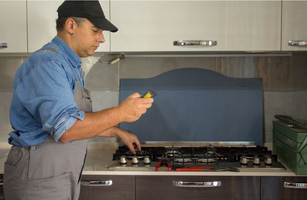 A repairman wearing a cap and overalls inspects a gas stove in a kitchen, holding a tool in one hand and adjusting the stove with the other. Various tools are laid out on the countertop.