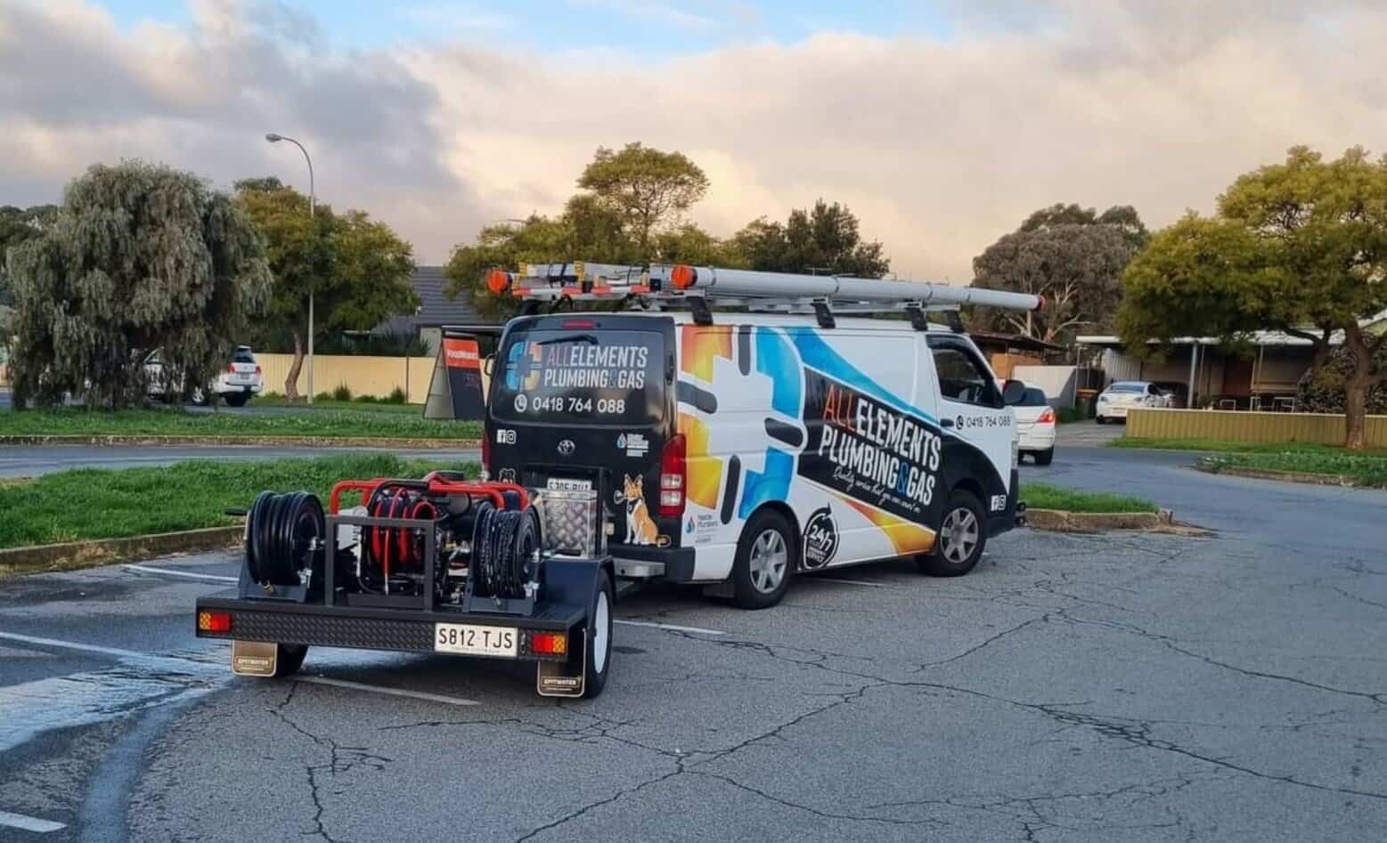 A plumber kneels on a suburban street, using equipment to inspect a drain. A branded plumbing van is parked nearby, and houses and greenery are visible in the background.