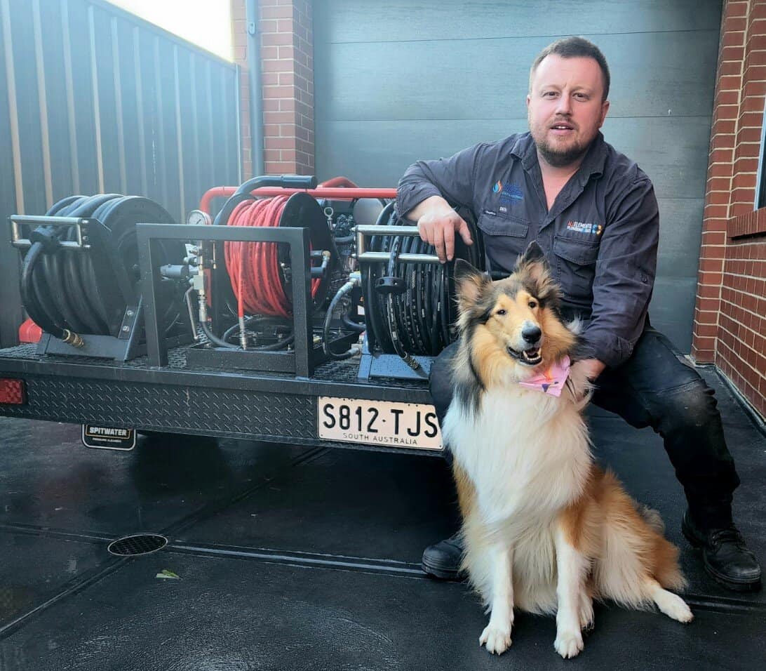 A man in work clothes kneels next to a collie dog