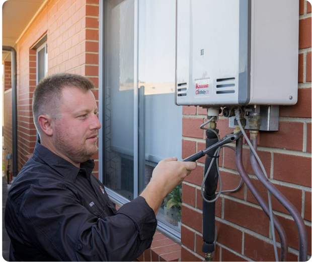 A man in a black shirt uses a wrench to work on an outdoor tankless water heater mounted on a brick wall next to a window.