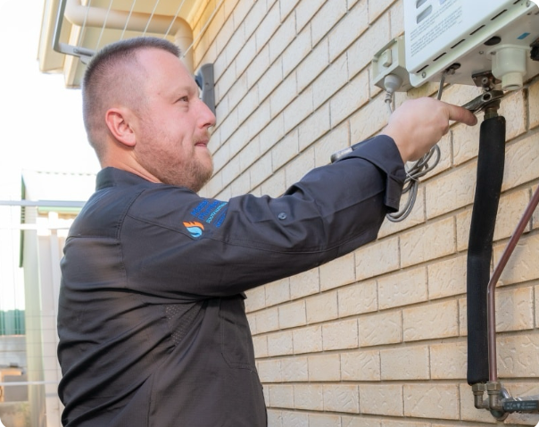 A technician wearing a dark uniform adjusts a device mounted on an exterior brick wall, working with pipes and cables connected to it.