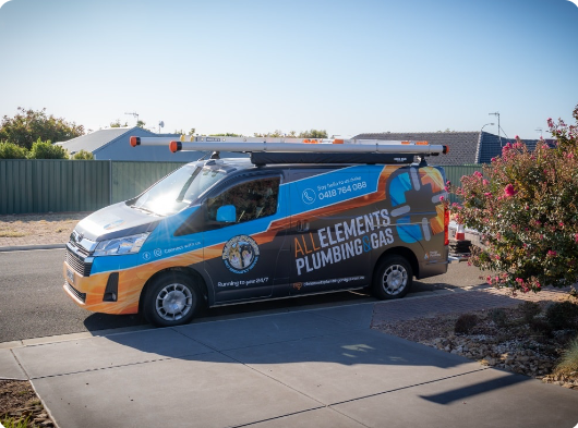 A van with "All Elements Plumbing + Gas" branding is parked on a residential street. The van has a roof rack and company contact details displayed on the side. Trees and houses are visible in the background.