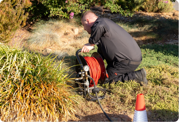 A person in black clothing kneels on the ground outdoors, inspecting or adjusting a hose reel in a garden area, with a traffic cone nearby and greenery in the background.