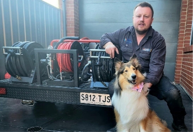 A man in work clothes kneels beside a Collie dog with a pink bow on its collar, in front of a trailer with hoses and equipment, next to a brick wall.