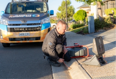 A plumber kneels on a suburban street, using equipment to inspect a drain. A branded plumbing van is parked nearby, and houses and greenery are visible in the background.