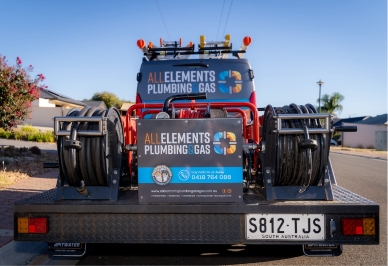 A plumbing service truck loaded with hoses and equipment shows branding for "All Elements Plumbing & Gas" and a South Australia license plate, parked on a suburban street.