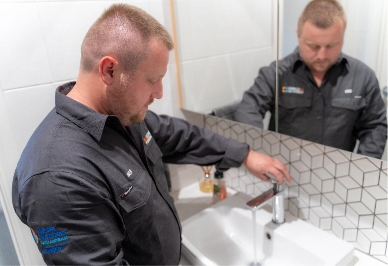A man in a dark work shirt stands in a bathroom adjusting a faucet on a white sink, with a mirror reflecting his image and geometric tiles on the wall.