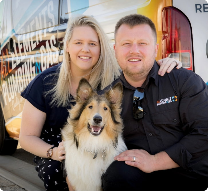A smiling woman and man kneel beside a happy Collie dog in front of a van with "ELEMENTS PLUMBING & GAS" written on it. The man wears a branded work shirt. All three look at the camera.