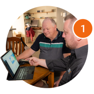 Two men sit at a table looking at a laptop screen together, with one man pointing at the screen. A large orange circle with the number 1 is overlaid in the upper right corner.