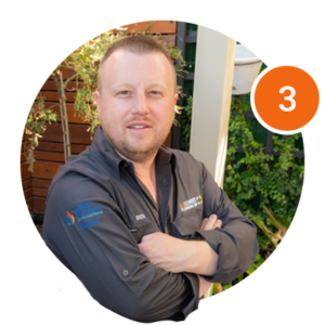 Man with short hair and a beard, wearing a dark gray shirt with embroidered logos, stands with arms crossed outdoors. Orange circle with the number 3 is in the top right corner of the image.