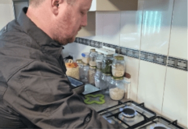 A man in a dark shirt stands beside a stove in a kitchen, preparing food. Various jars and containers are arranged on the countertop in the background.