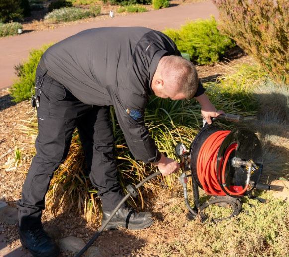 A plumber in a dark uniform stands next to a colorful service van with "The Calile Plumber" logo, holding a red tool bag. The van has contact details and plumbing equipment visible inside.