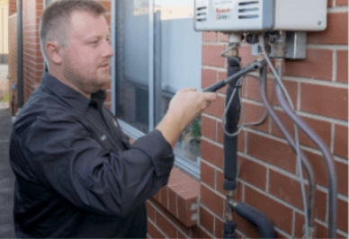 A man wearing a dark shirt uses a wrench to work on outdoor piping connected to a wall-mounted tankless water heater attached to a brick wall.