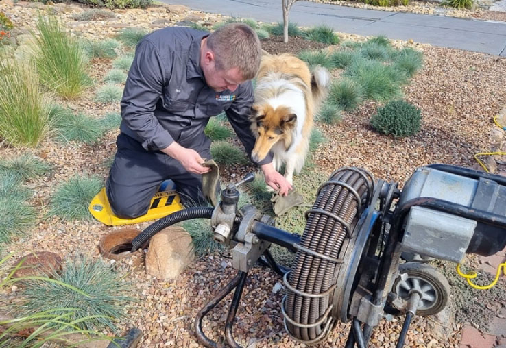 A man in a dark shirt is servicing or inspecting a gas water heater mounted on the exterior brick wall of a house. He is using a tool to adjust or check the pipes connected to the unit.