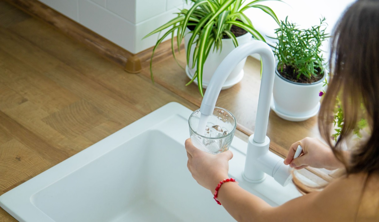 A person fills a glass with water from a white kitchen faucet, surrounded by green potted plants on a wooden countertop next to a white sink.