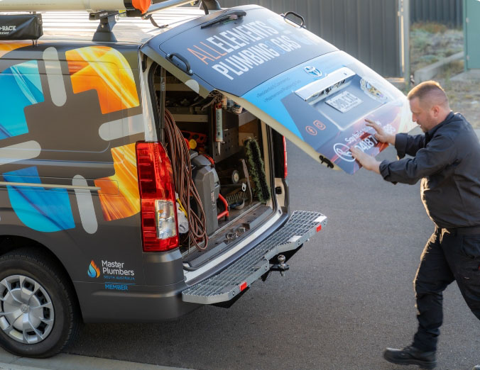 A plumber wearing dark clothes closes the rear door of a van labeled "Allegiance Plumbing Gas." The van’s back is open, revealing tools and equipment inside. The van displays a colorful logo and "Master Plumbers Member.