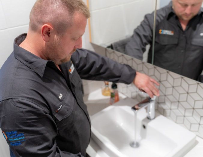 A plumber in a dark uniform turns on a modern bathroom faucet, checking the water flow at a white sink with patterned tile backsplash. His reflection is visible in the mirror above the sink.
