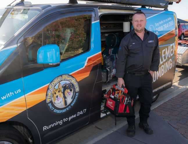A plumber in a dark uniform stands beside a colorful service van with “The Coffey Plumbers” logo, holding a tool bag. The van’s side door is open, showing plumbing tools and equipment inside.