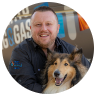 A smiling man with short hair and a beard sits next to a happy collie dog. Both are facing the camera. There is a blurry background with colorful signs and text.