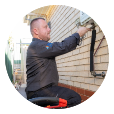 A man in a dark uniform works on an exterior utility meter attached to a brick wall, using tools to perform maintenance or repairs.