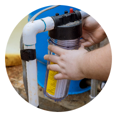 A person’s hands are installing or servicing a clear water filter canister attached to white PVC pipes, with a blue tank visible in the background.