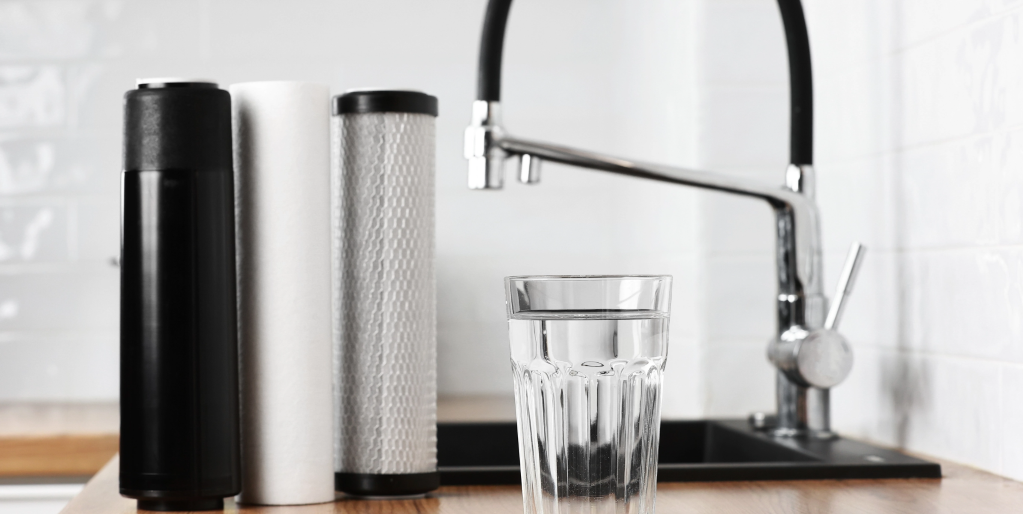 A glass of clear water sits on a kitchen counter beside three water filter cartridges and a modern faucet, with a black sink and white tiled wall in the background.